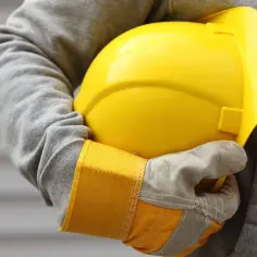 Osoba w szarej bluzie i rękawicach trzyma żółty kask ochronny.