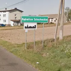 Znak drogowy z napisem "Rębielice Szlacheckie" - to najdłuższa nazwa miejscowości w Polsce. W tle domy i pola.