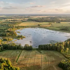 Krajobraz z lotu ptaka: jezioro, lasy i pola uprawne. Słońce zachodzi, rzucając złote światło na ziemię, jakby opowiadało starą historię w języku kaszubskim.